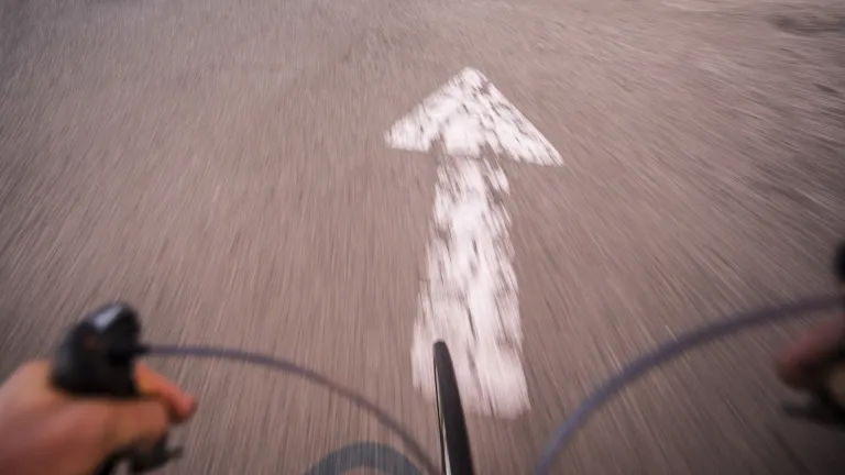 First-person view of a person on a bicycle looking down at a paved path with a white arrow on it.